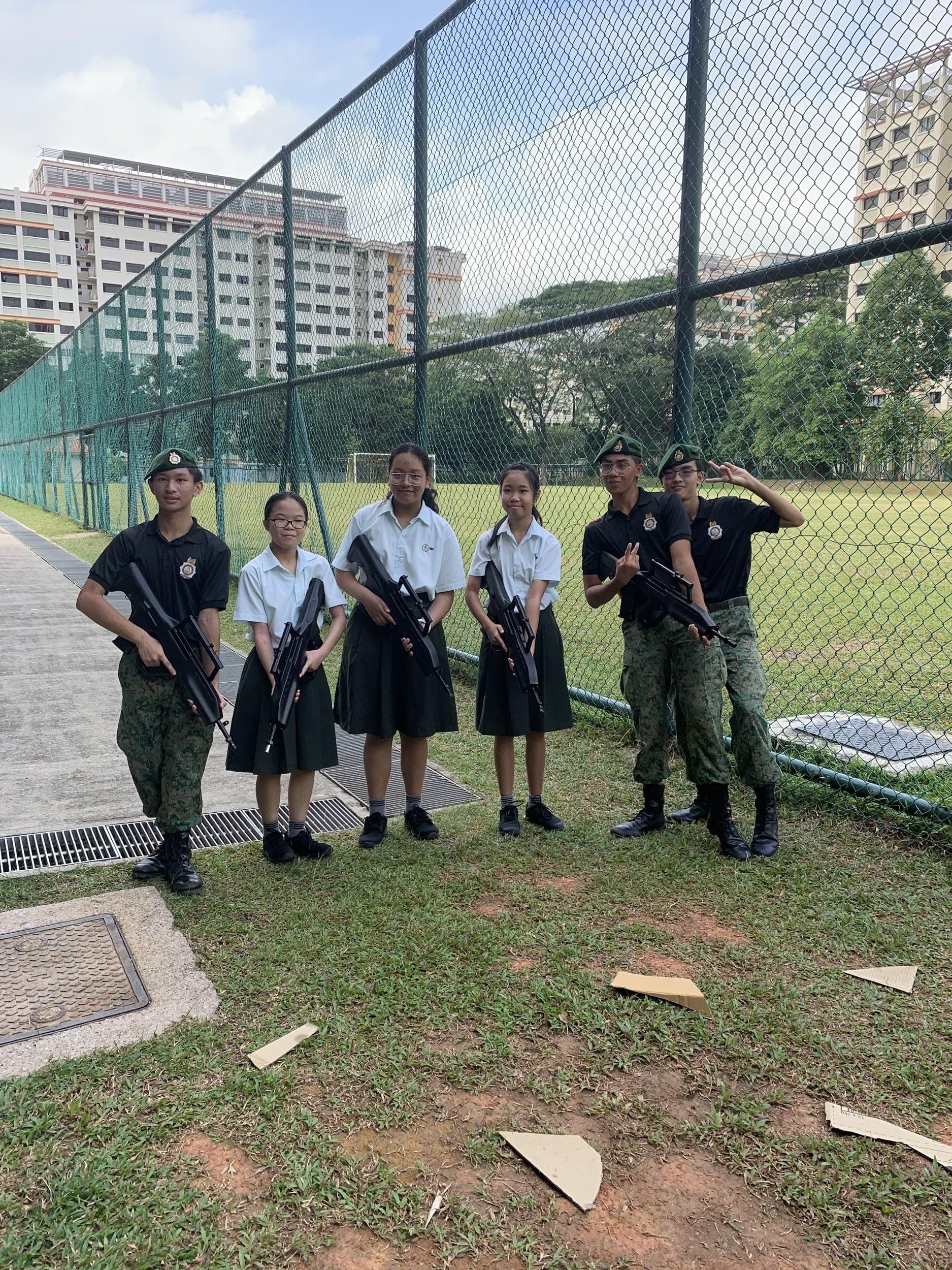 Six young students in school and military uniforms hold rifles outdoors near a green chain-link fence and field.